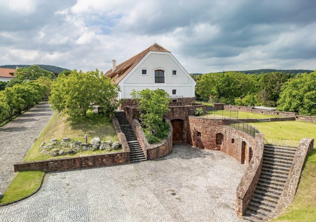 White, gable-roofed building atop stone fortress walls with staircases, surrounded by green trees and a cobblestone courtyard amid a cloudy sky.