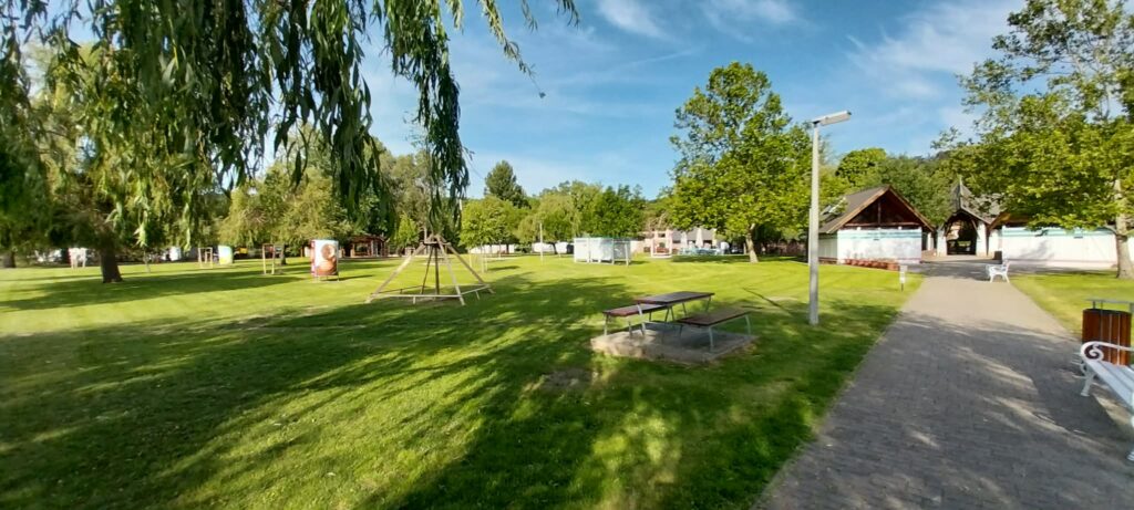 Sunlit park with green grass, mature trees, and a paved path; benches and wooden pavilions in the background.