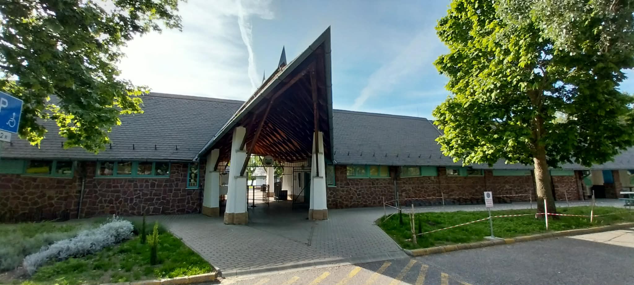 Public building with a tall, triangular-roof entrance supported by white columns, stone walls, and green-framed windows, shaded by trees on both sides.