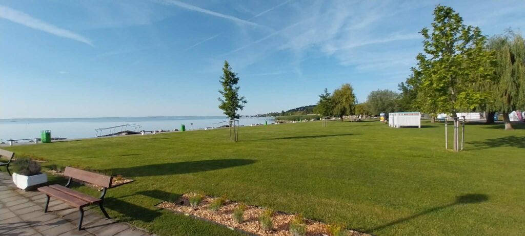 Sunlit lakeside park with green lawn, benches, and trees along a calm bay by the water's edge.