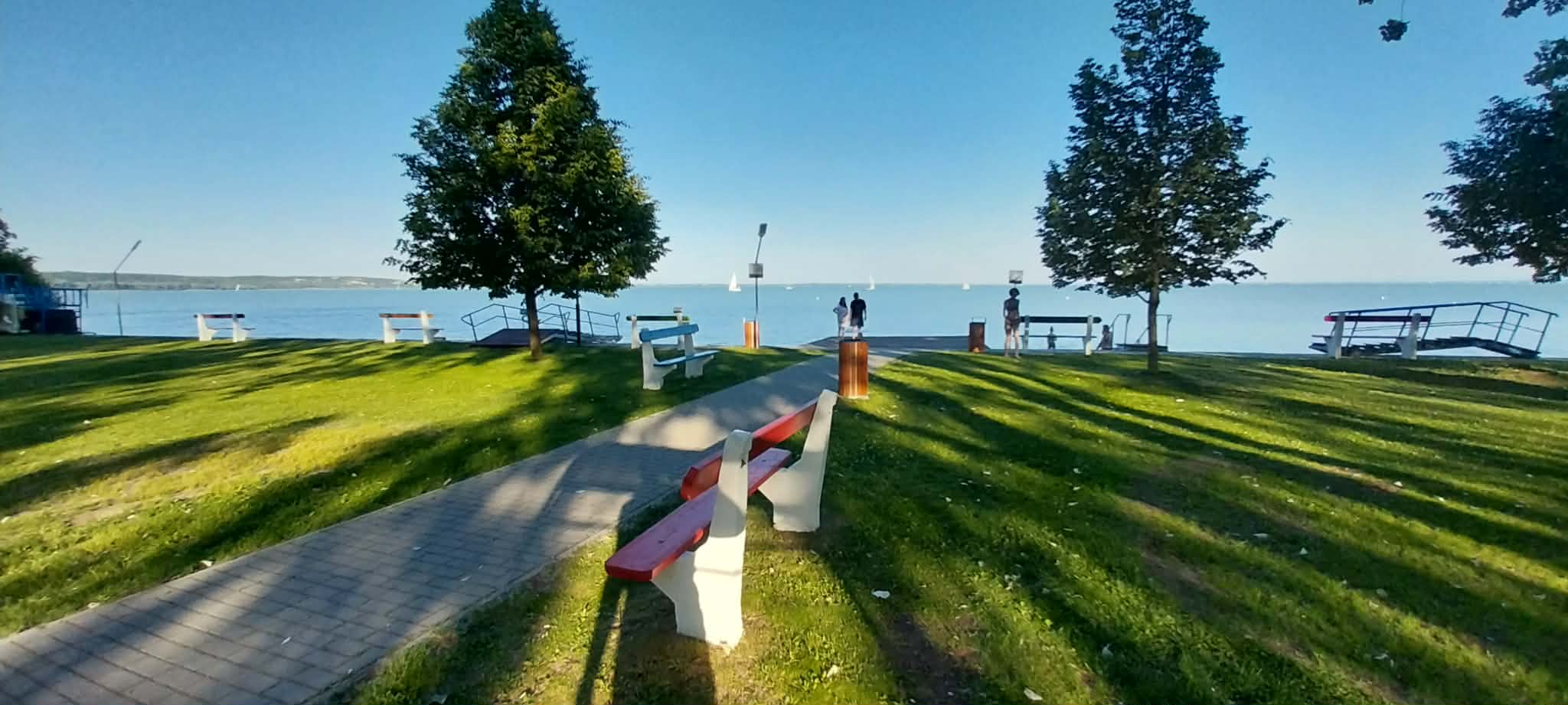 Lakeside park with green grass, benches, and a paved path; people near the water with sailboats on the lake.