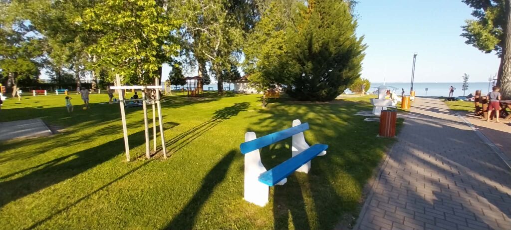 Sunny seaside park with a blue-and-white bench in the foreground, tall trees, and people along a promenade by the water.