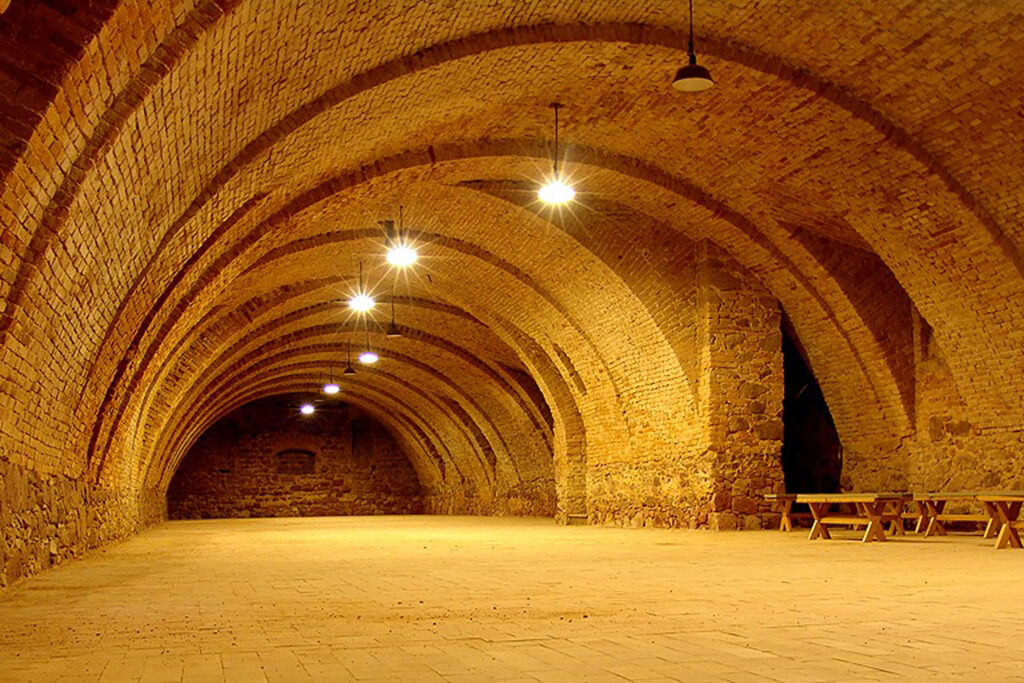 Long brick arch tunnel illuminated by hanging lamps, empty except for wooden benches on the right side.