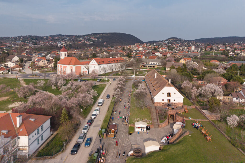 Aerial view of a European town square with a pink church, red roofs, blooming trees, and people at an outdoor market or festival.