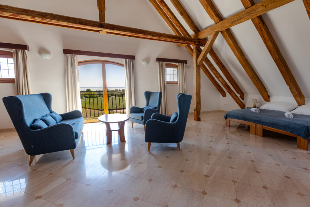 Attic living area with blue upholstered chairs around a round table, exposed wooden beams, and a bed on the right. Balcony doors overlook a lawn and water view.