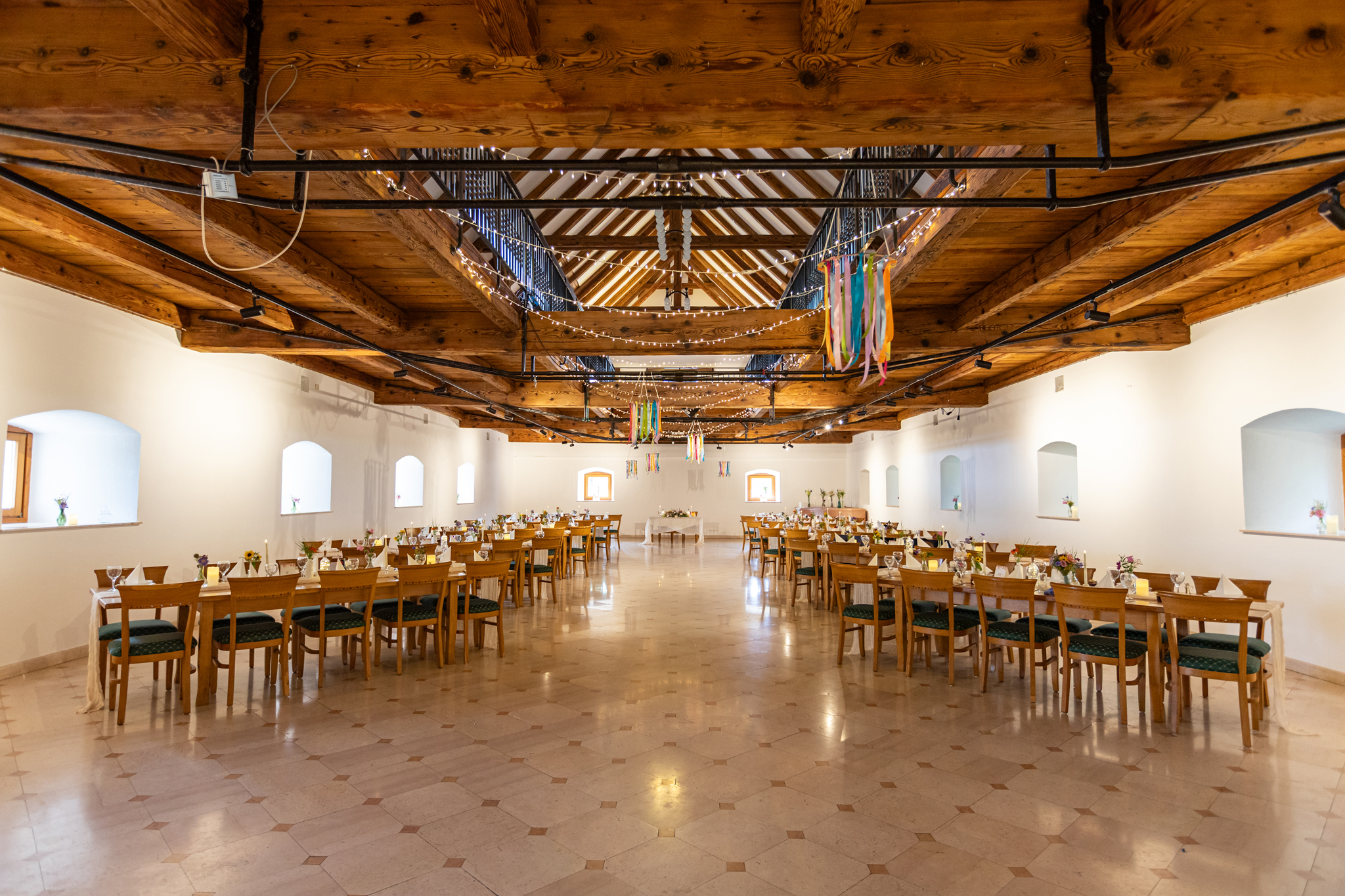 Banquet hall with exposed wooden beams, string lights, and long tables set for an event under a high ceiling.