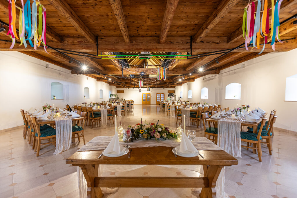 Rustic banquet hall with exposed wooden beams, long tables, and colorful ribbon garlands overhead for a festive event.