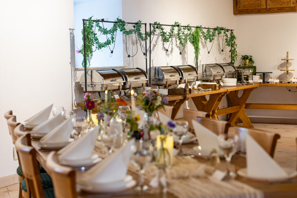 Buffet line with stainless steel chafing dishes on a wooden table, green ivy garlands overhead in a bright dining room