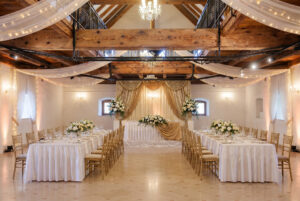 Wedding reception hall with long white-draped tables, gold chairs, and floral centerpieces under chandeliers in a rustic venue.