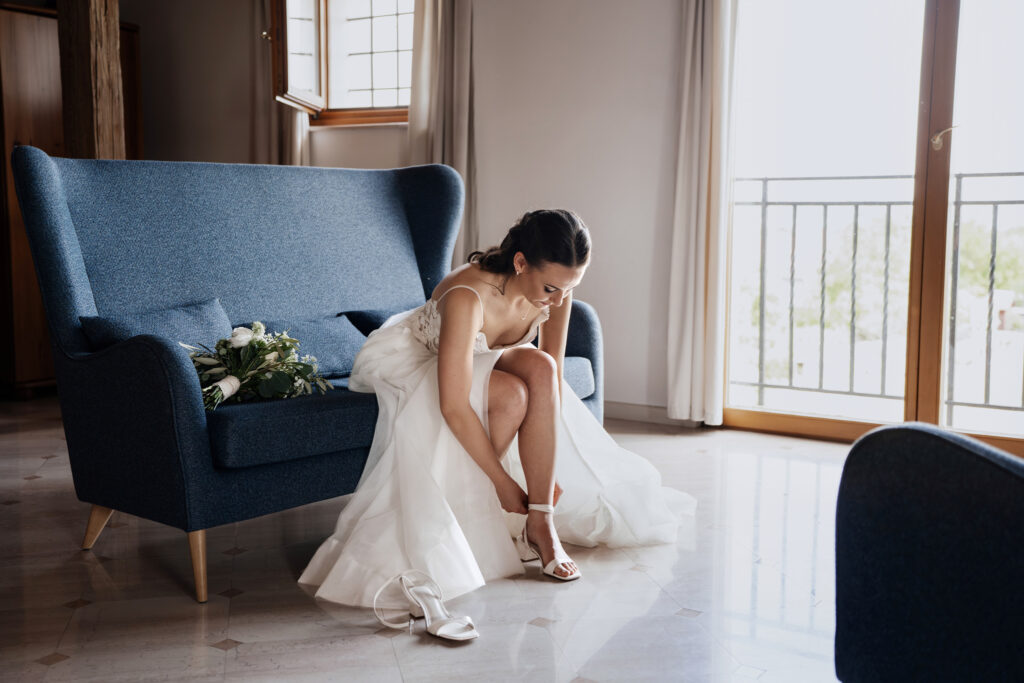 Bride in a white wedding dress sits on a blue sofa, tying her white heel in a sunlit room with a bouquet nearby.