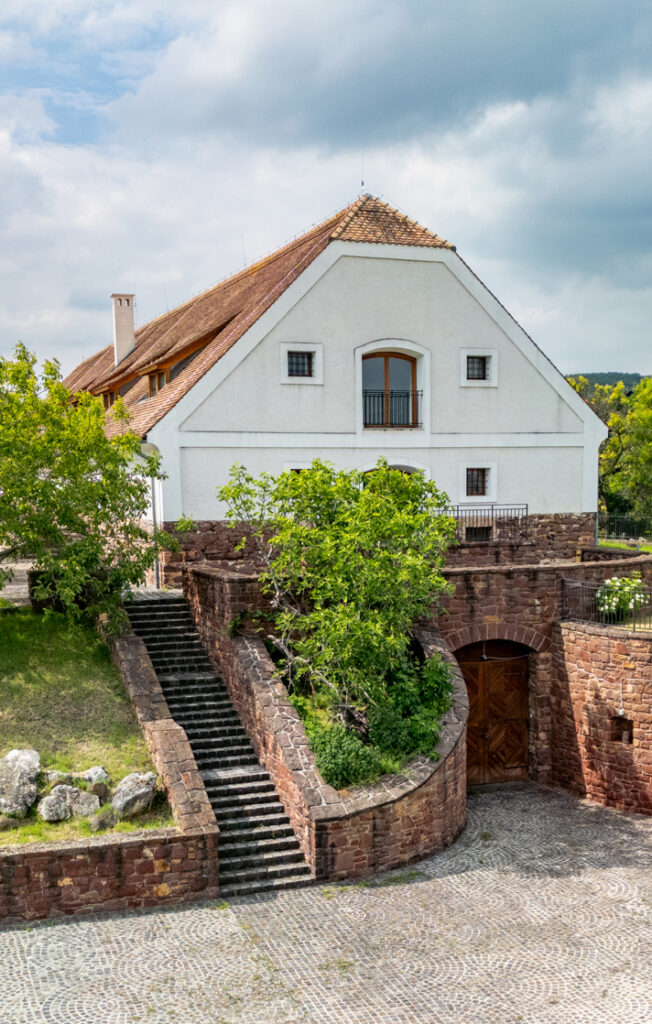 White house with a steep red-tiled roof perched on a stone terrace, accessible by a grand stone staircase and surrounded by green trees under a cloudy sky.