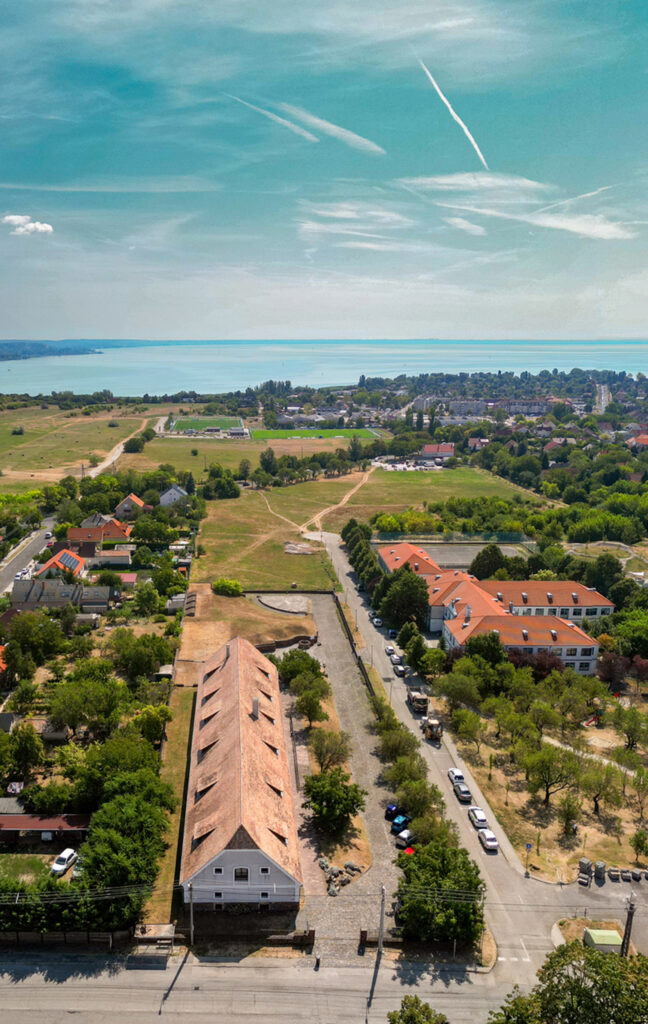 Aerial view of a town with a long brick warehouse along a tree-lined street, surrounding houses, and a distant river into the horizon on a sunny day.