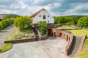 White two-story house with a red-tiled gable roof perched on a stone fortress, surrounded by trees and a cobblestone courtyard under a cloudy sky.