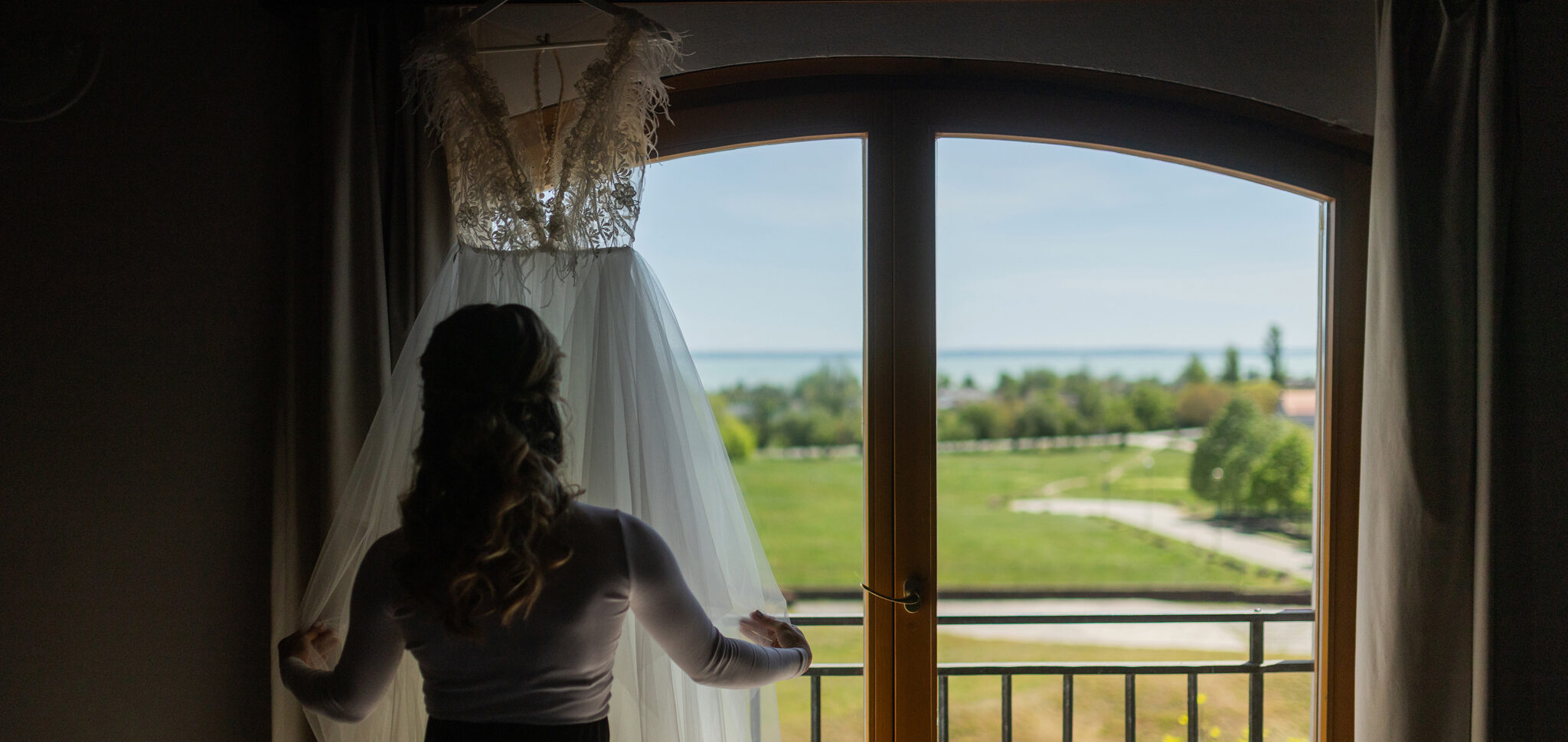 Bride seen from behind, adjusting a lace veil in a white wedding gown by a large window, with a green lawn and lake visible outside.