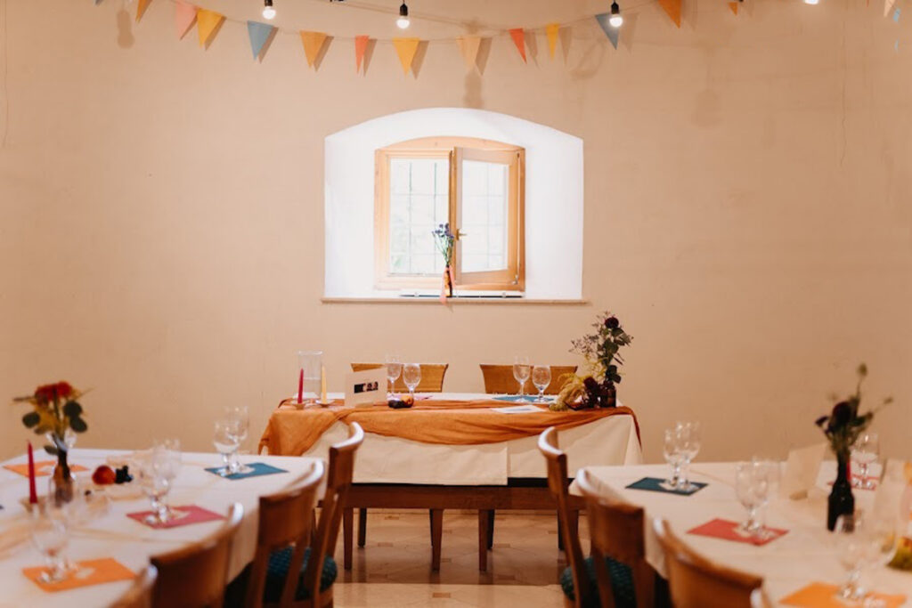 A festively set dining room with long tables, orange tablecloths, folded napkins, and floral centerpieces under colorful pennant bunting tied across the ceiling.