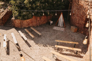Open-air stone courtyard with string lights, wooden benches, and a patterned rug along a cobblestone ground.