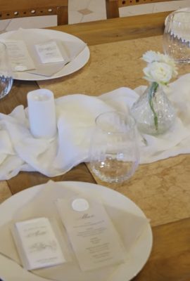 White table setting with a plate, napkin, menu card, and a small white candle, plus a white flower in a glass vase centerpiece.