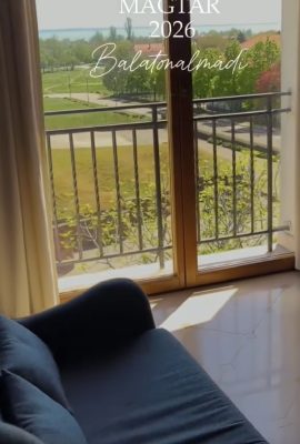 Living room scene with a glass balcony door and railing; sunlight on the floor and a park view, with overlay text 'MAGTÁR 2026 Balatonalmádi'.