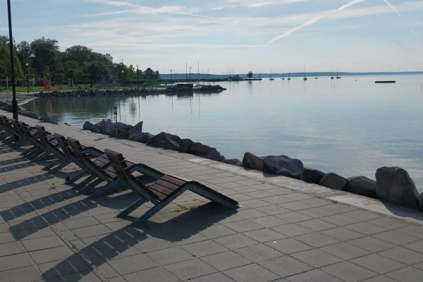 Row of empty leaning deck chairs along a lakeside walkway beside calm water and a rocky edge.