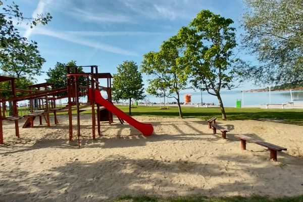 Wooden playground with a red slide and a green slide on sandy ground beside a calm lake, shaded by trees with benches nearby.